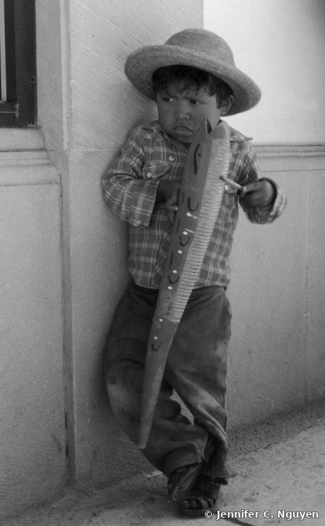 Original black-and-white archival photograph of a young boy wearing a hat, leaning against a wall and holding a decorative wooden tool