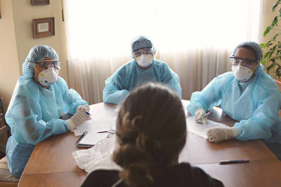 Three healthcare workers in blue PPE gowns, masks, and face shields seated at a table reviewing paperwork with a patient during an in-home visit