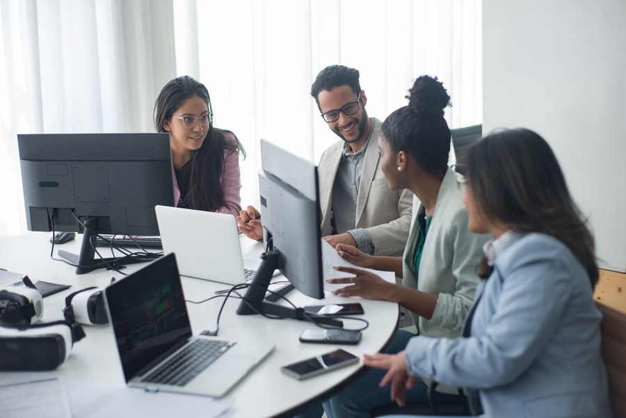 Four colleagues gathered around a bright desk with dual monitors, laptops, and VR headsets, smiling and discussing a document together in a modern office