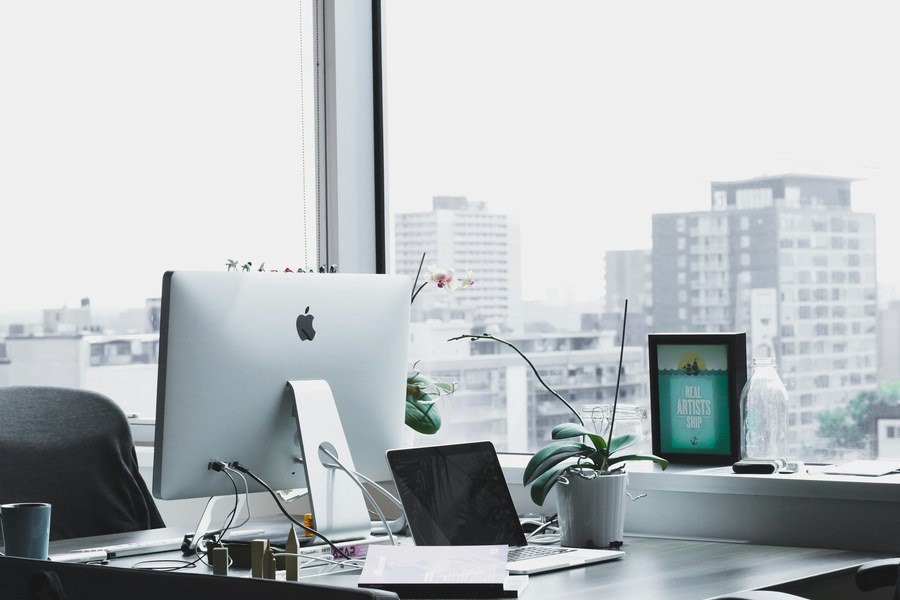 Clean minimalist desk setup with an iMac, laptop, orchid plant, and a framed Real Artists Ship poster against a city window view