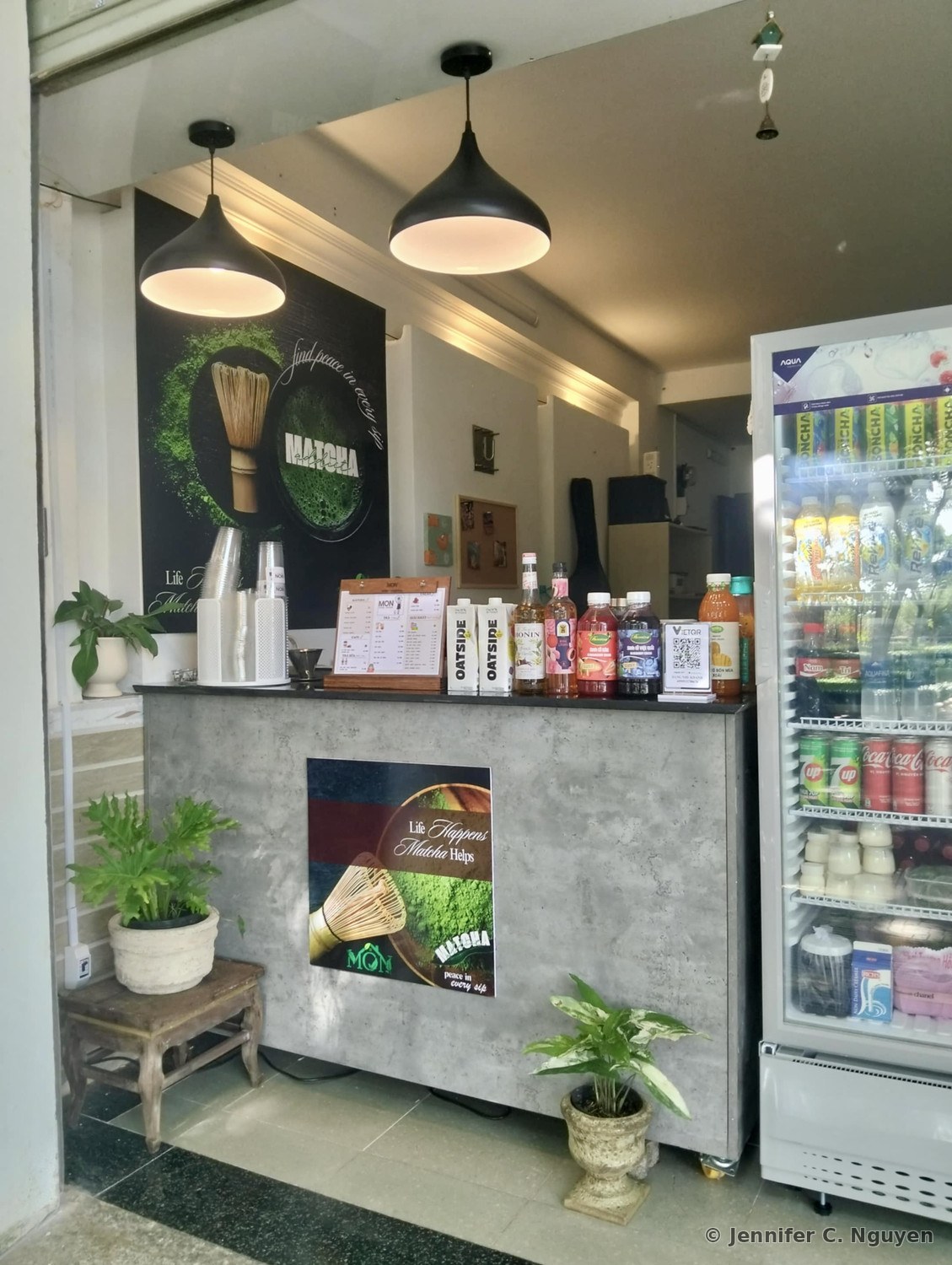 Both matcha posters printed and mounted in person at Mon Cafe's counter, the brown three-panel poster hanging on the wall above the drink station and the dark centered-logo poster mounted on the concrete counter face, surrounded by potted plants, a drink menu, and a beverage refrigerator