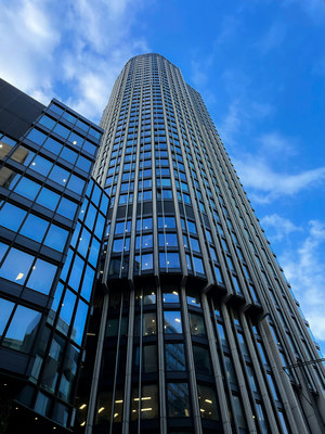 Upward view of a tall curved gray skyscraper with vertical ribbed columns against a blue sky with scattered clouds, a glass office building visible to the left