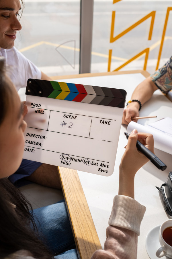 Close-up of a film clapperboard reading Scene number 2 held up during a video production planning session, with two people writing notes nearby